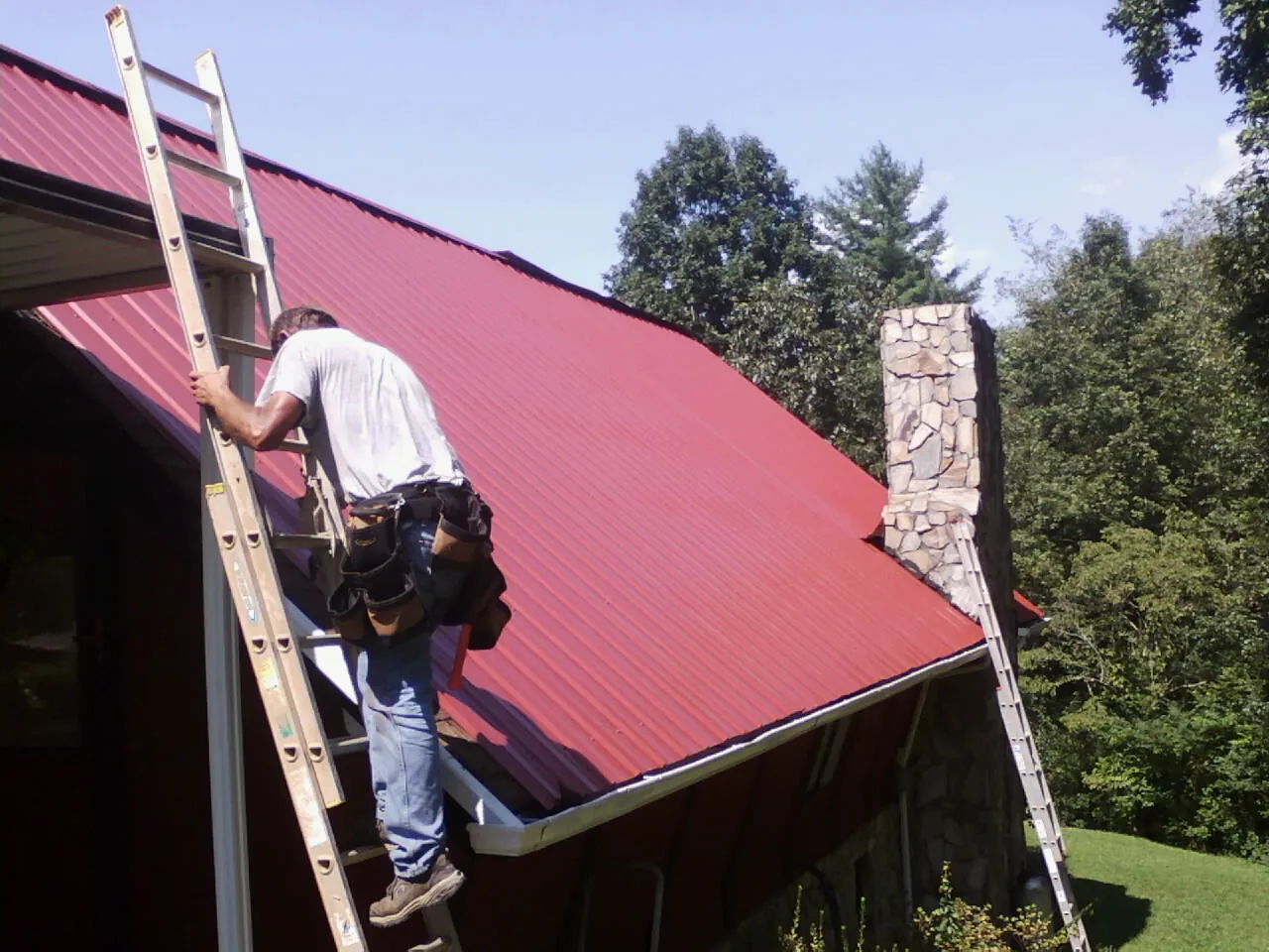 Roofer on a ladder beside a red metal roof and stone chimney — Soffit & Fascia Repair by Roofing Brothers in Loch Sheldrake